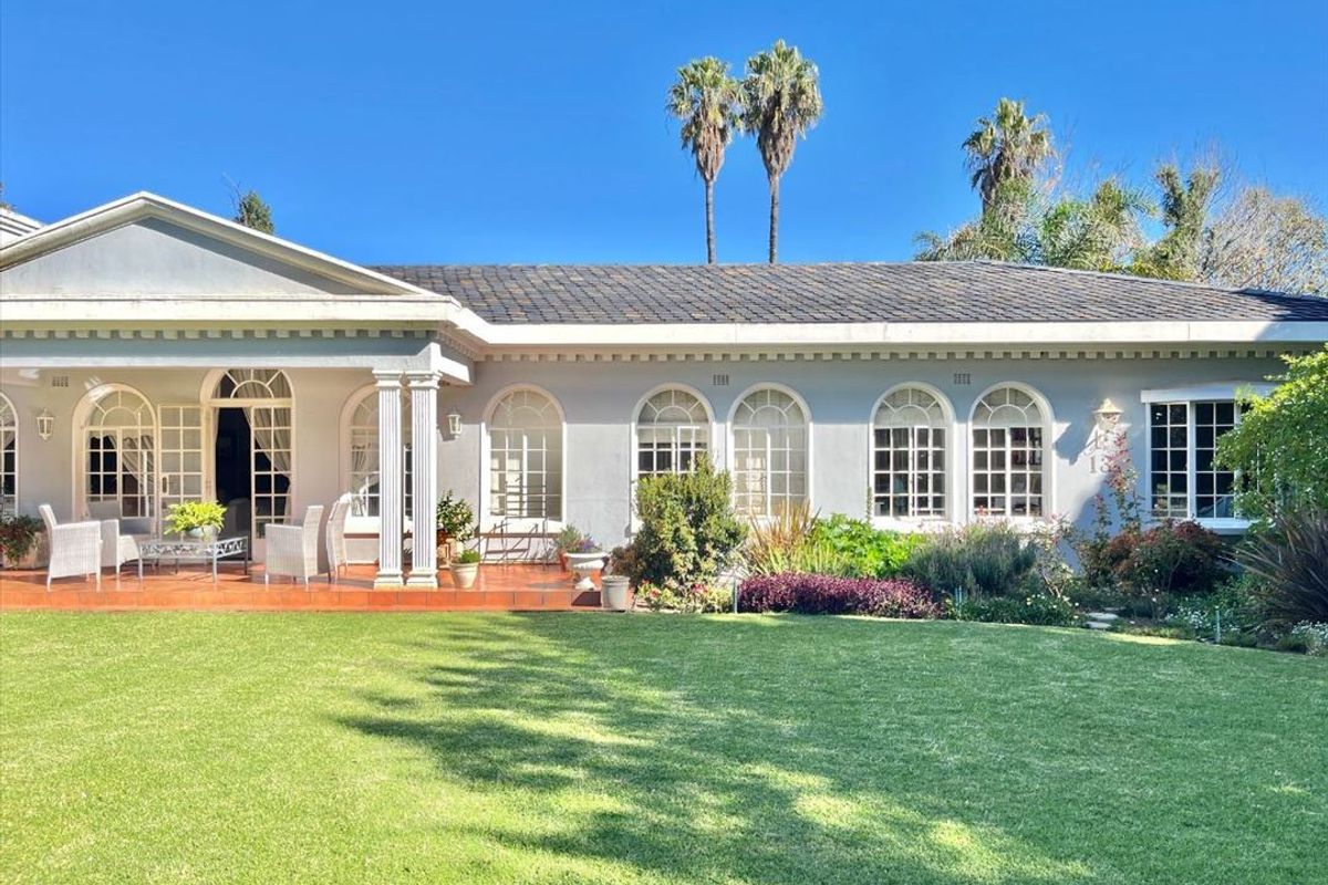 A well-maintained rear exterior view featuring a spacious lawn, a covered tiled patio with classical columns, and elegant arched windows.