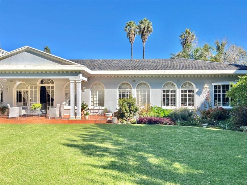 A well-maintained rear exterior view featuring a spacious lawn, a covered tiled patio with classical columns, and elegant arched windows.