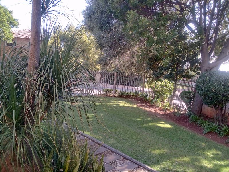 A well-maintained private garden featuring a lush lawn, mature trees, and secure metal perimeter fencing with a paved walkway.