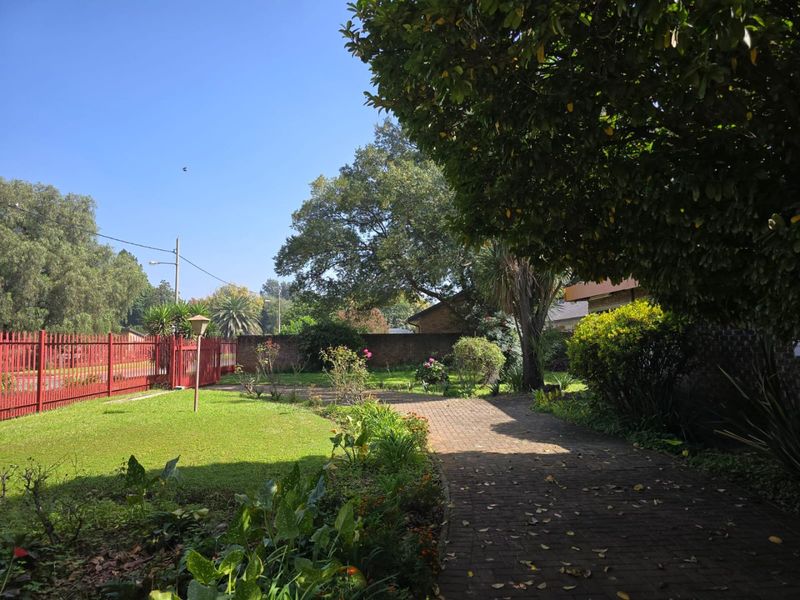 A well-established garden featuring a brick-paved driveway, lush lawn, mature trees, and secure perimeter fencing.