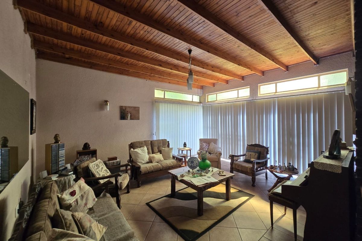 Spacious living room featuring a high exposed timber beam ceiling, tiled flooring, and large windows with vertical blinds allowing for natural light.
