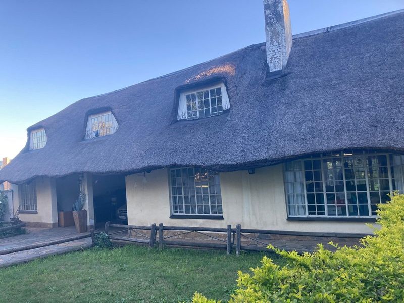 Front elevation of a traditional cottage featuring a thatched roof, dormer windows, and stone paving, set within a landscaped garden area.
