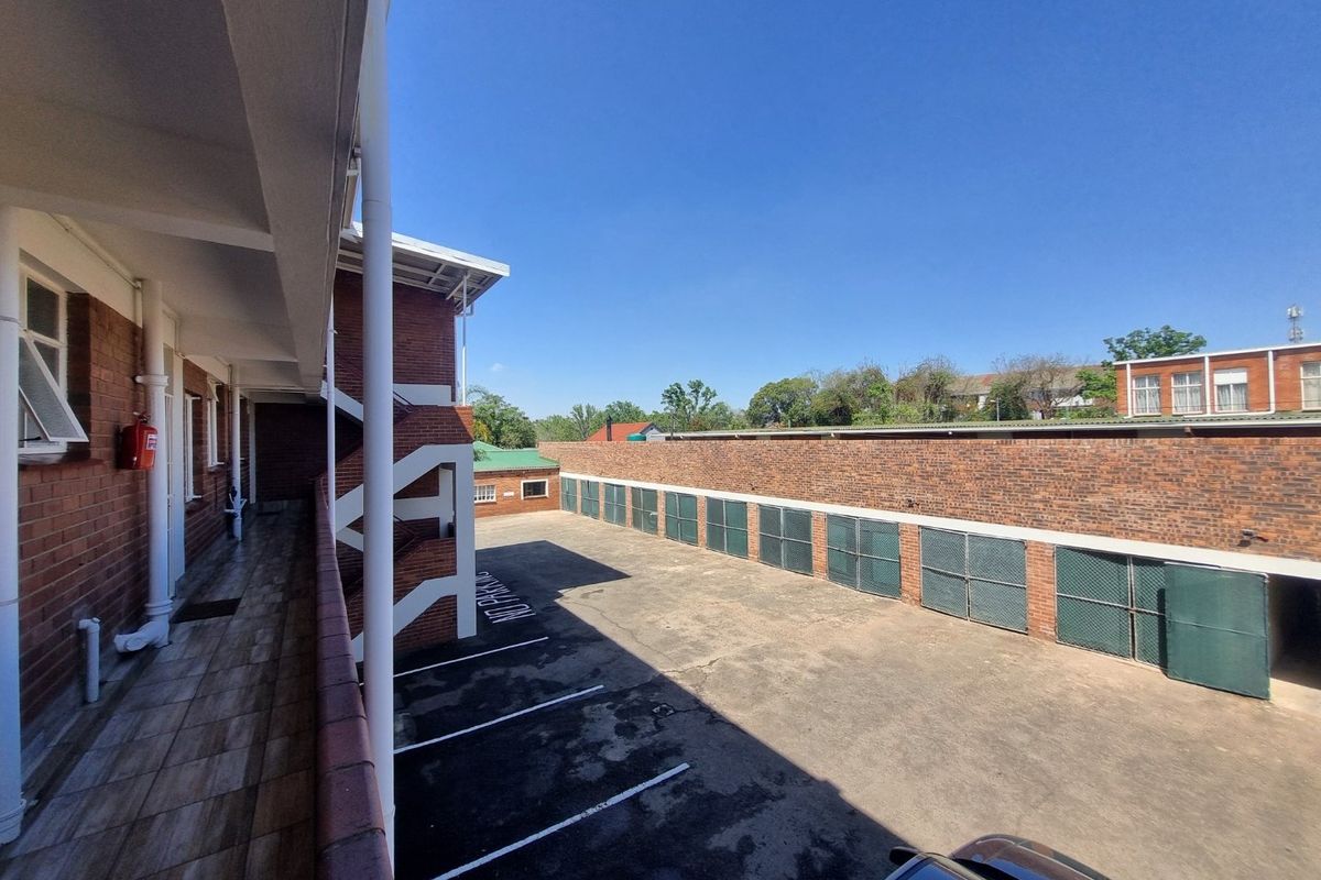 An exterior view of the apartment complex rear area, featuring a paved parking lot, secure metal storage units, and a covered tiled walkway.