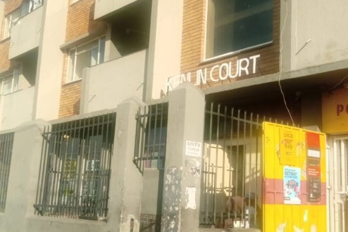 Street-level view of the Kenin Court apartment building facade, featuring brick exterior, security fencing, and a concrete boundary wall.
