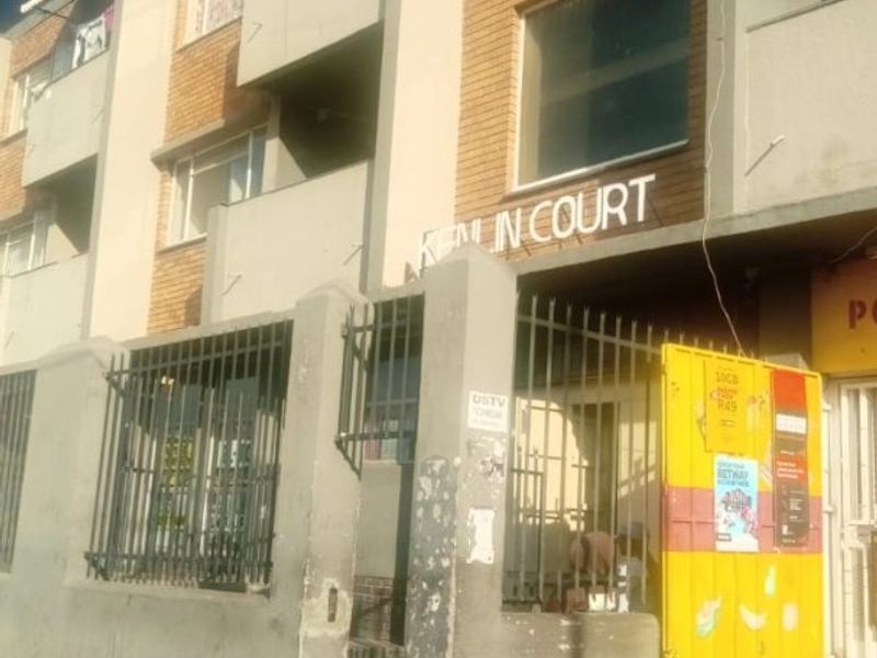 Street-level view of the Kenin Court apartment building facade, featuring brick exterior, security fencing, and a concrete boundary wall.