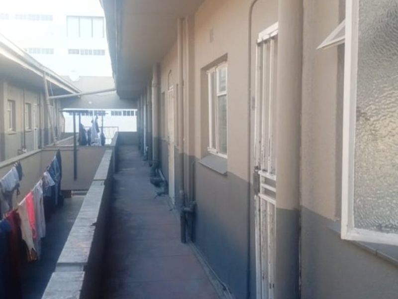 An exterior apartment walkway featuring concrete flooring, security gates, and exposed plumbing pipes along the building facade.