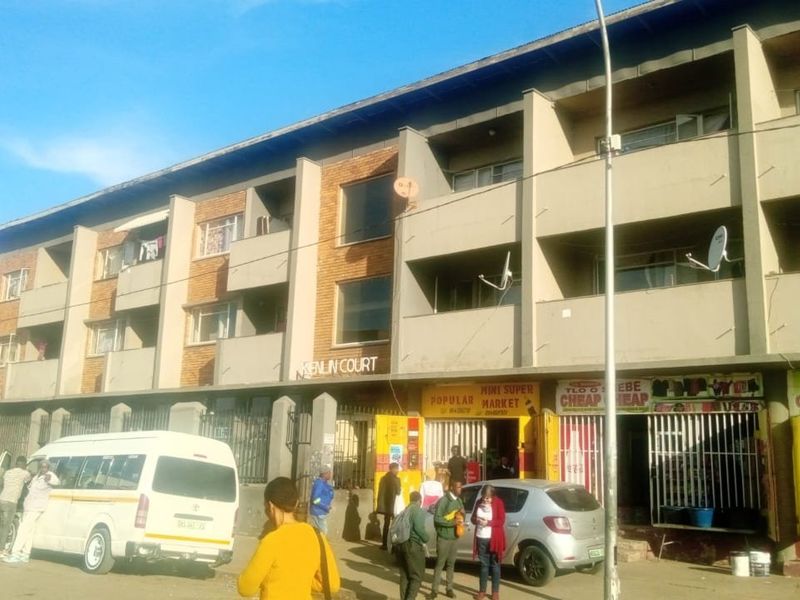 Street-level view of Kenlin Court, a multi-story apartment building featuring residential balconies above ground-floor commercial retail spaces.