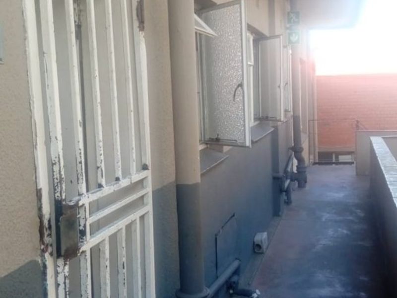 An exterior apartment balcony featuring a security gate, exposed plumbing, and concrete flooring, showing signs of wear and requiring maintenance.