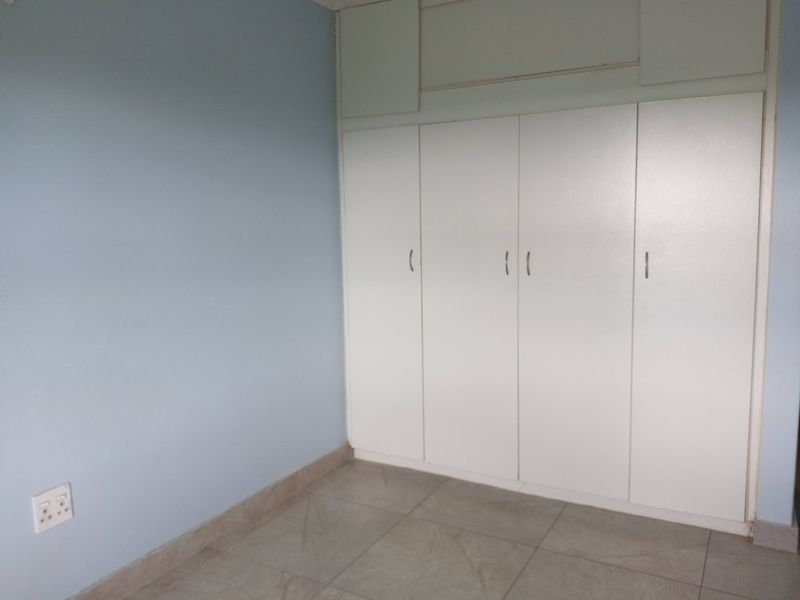 A clean, empty bedroom featuring neutral tiled flooring, light blue walls, and ample built-in cupboard storage with overhead cabinets.