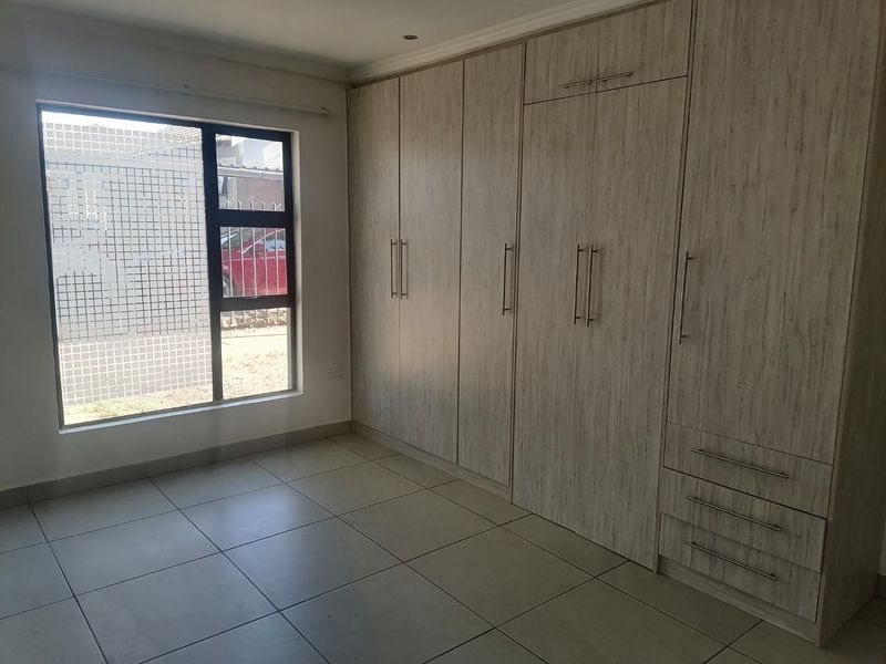 A clean, empty bedroom featuring modern light-wood built-in cupboards, neutral floor tiles, and a window with security bars.