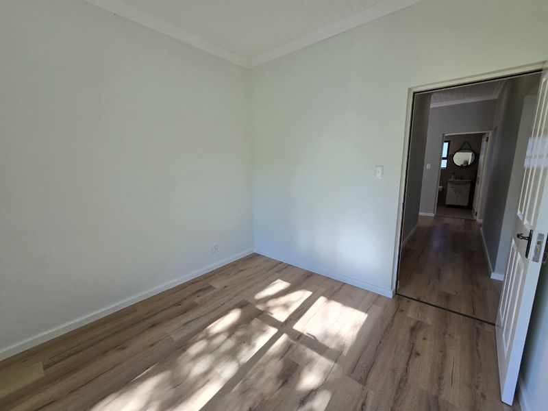 A bright, empty bedroom featuring modern laminate flooring, neutral walls, and a doorway leading to an adjacent hallway and bathroom.