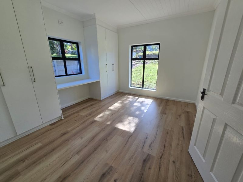 A bright, empty bedroom featuring modern laminate flooring, built-in cupboards, a built-in desk, and large windows with black aluminium frames.