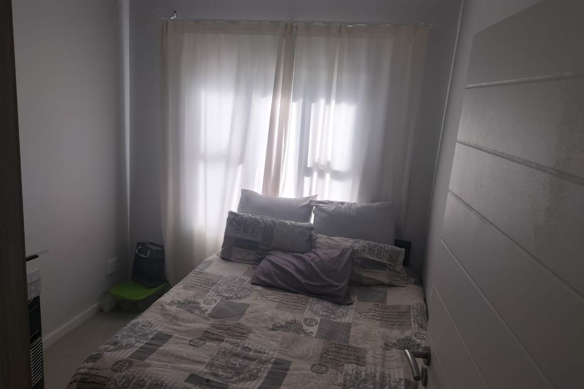 A compact bedroom featuring tiled flooring and neutral wall finishes, illuminated by natural light through a window with sheer curtains.