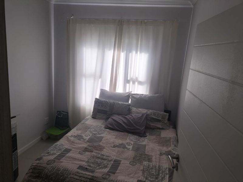 A compact bedroom featuring tiled flooring and neutral wall finishes, illuminated by natural light through a window with sheer curtains.