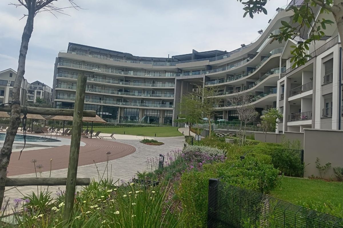 A view of the rear apartment complex facade featuring landscaped gardens, a swimming pool area, and modern balconies with glass balustrades.