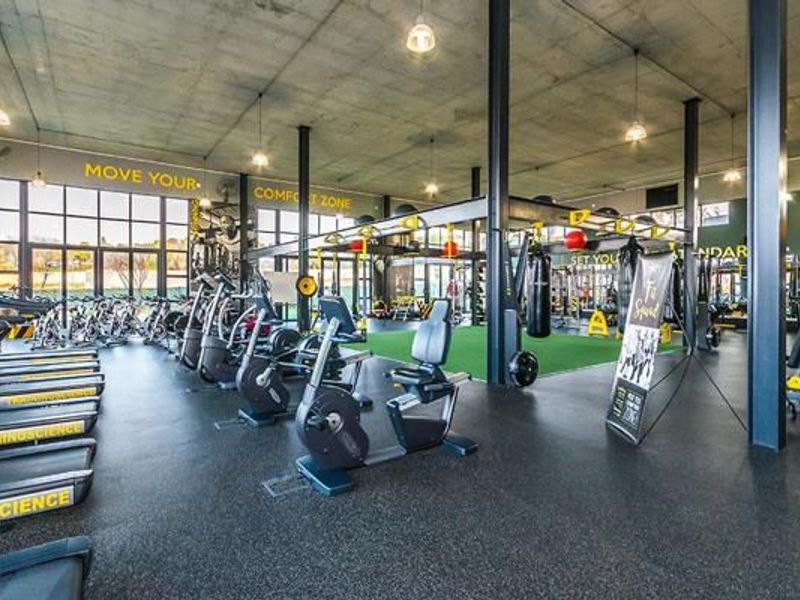 A spacious, modern gym facility featuring exposed concrete ceilings, steel structural columns, rubberized flooring, and an integrated artificial turf training area.