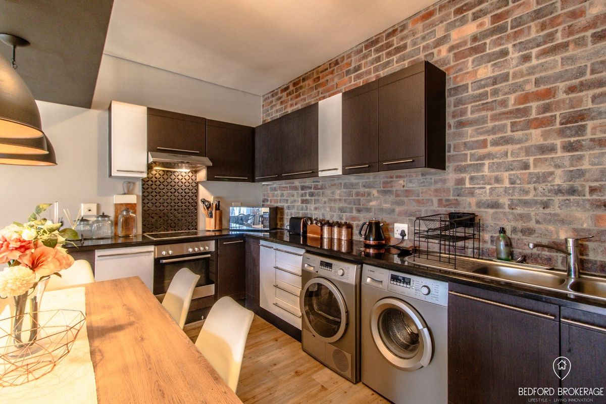 Modern kitchen featuring exposed brick walls, dark cabinetry, integrated appliances, and a functional layout with a stainless steel sink and built-in hob.