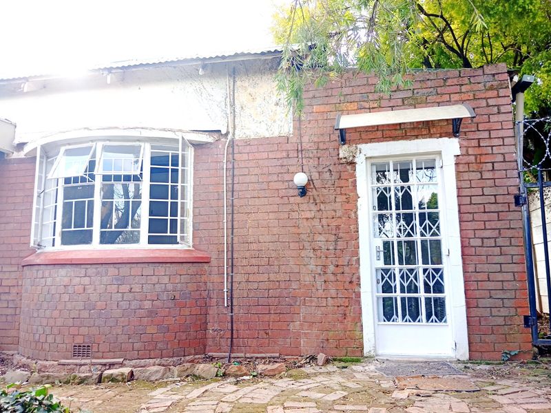 Rear exterior view of a brick house featuring a bay window, security gate, and paved area, showing signs of wear and maintenance requirements.