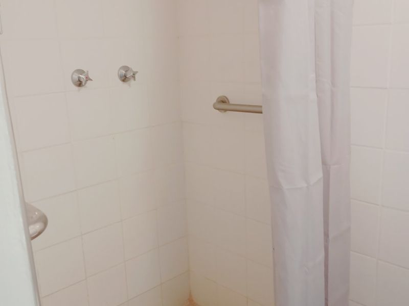 A functional shower enclosure featuring white wall tiles, mosaic floor tiling, a wall-mounted shower head, and a safety grab rail.