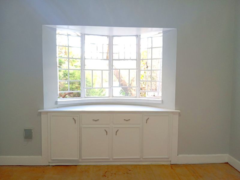 A bright bedroom featuring a classic bay window with integrated built-in storage cabinetry and original hardwood flooring.