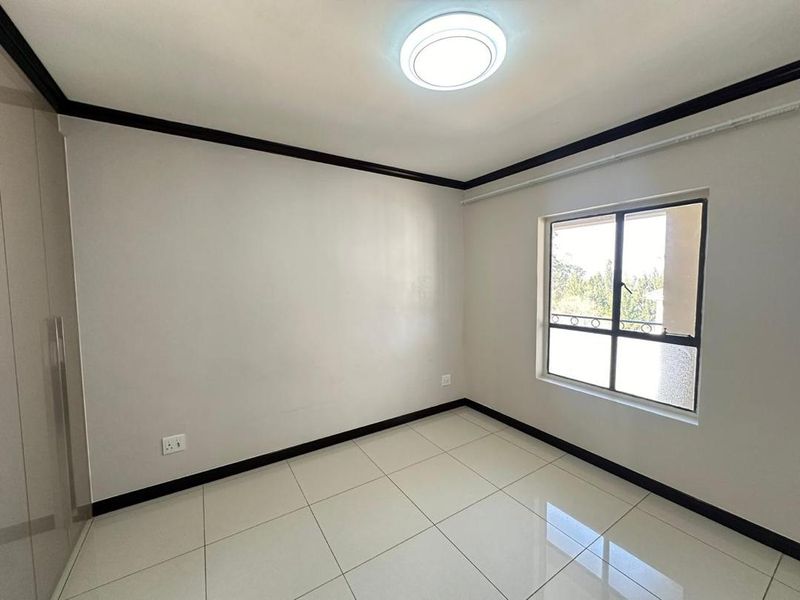 A clean, empty bedroom featuring neutral tiled flooring, built-in wardrobe storage, and a window with security bars, providing a bright and versatile living space.