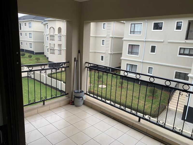 A clean, covered balcony featuring tiled flooring and a decorative wrought iron balustrade, overlooking the apartment complex grounds.