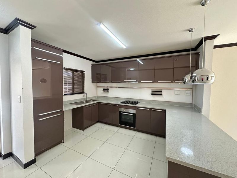 Modern kitchen featuring high-gloss brown cabinetry, stone countertops, built-in oven, gas hob, and tiled flooring in a clean, open-plan layout.