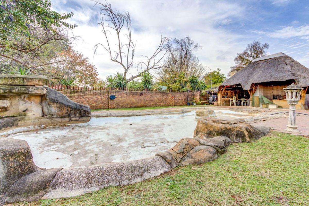 Outdoor pool area featuring a large rock-lined swimming pool, brick boundary wall, and a traditional thatched lapa entertainment space.