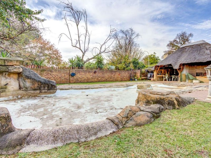 Outdoor pool area featuring a large rock-lined swimming pool, brick boundary wall, and a traditional thatched lapa entertainment space.