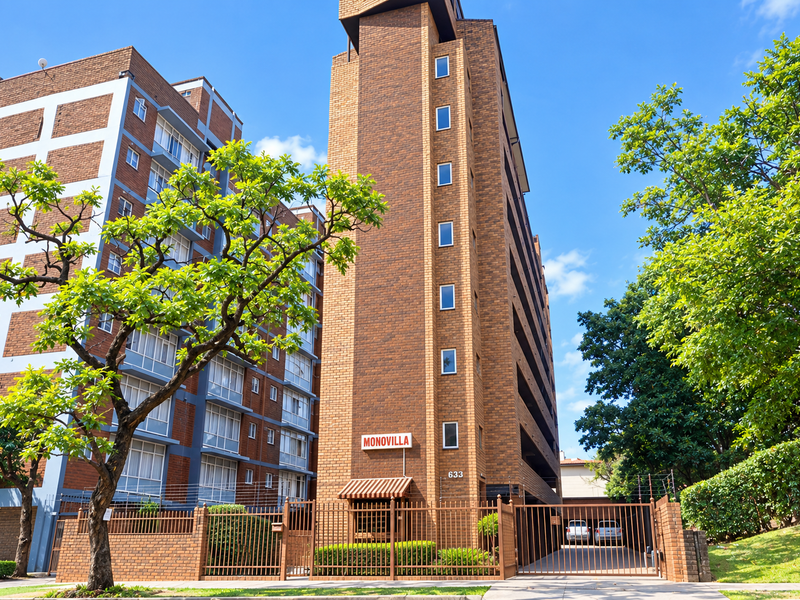 Exterior view of Monovilla apartment building featuring a brick facade, secure gated entrance, and established landscaping.