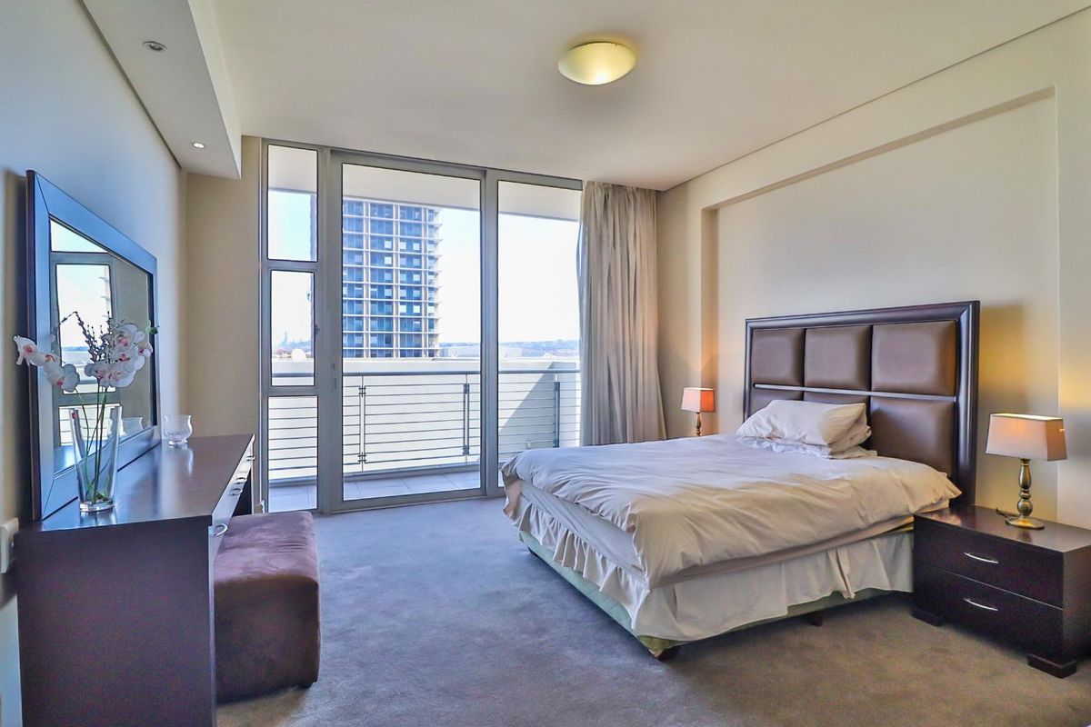 A spacious bedroom featuring neutral carpeting, recessed lighting, and a large sliding glass door providing access to a private balcony with urban views.