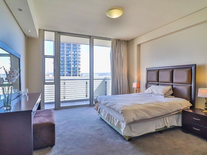 A spacious bedroom featuring neutral carpeting, recessed lighting, and a large sliding glass door providing access to a private balcony with urban views.