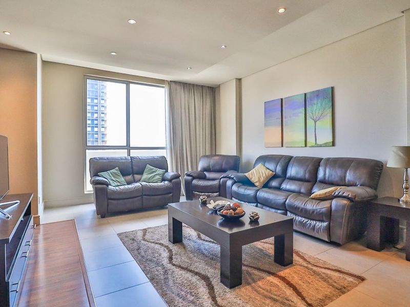 A well-lit living room featuring neutral tiled flooring, recessed ceiling lighting, and a large window providing natural light and urban views.