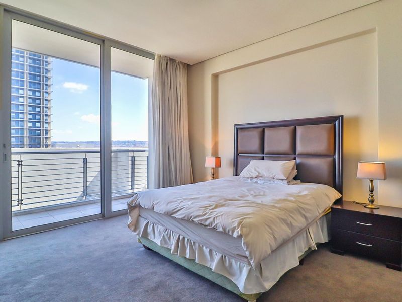A well-lit bedroom featuring floor-to-ceiling windows with access to a private balcony, neutral carpeting, and a modern recessed ceiling design.