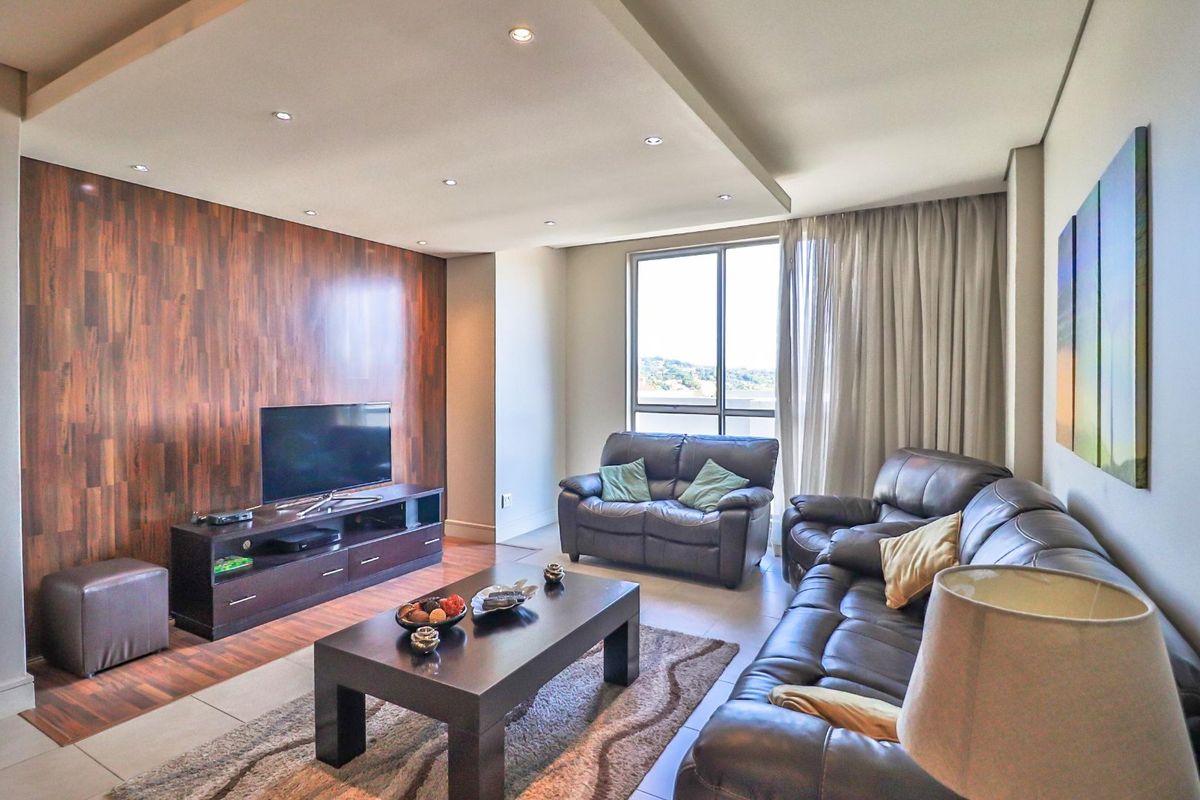 A well-maintained living room featuring a modern wood-paneled feature wall, recessed ceiling lighting, and large windows providing natural light.