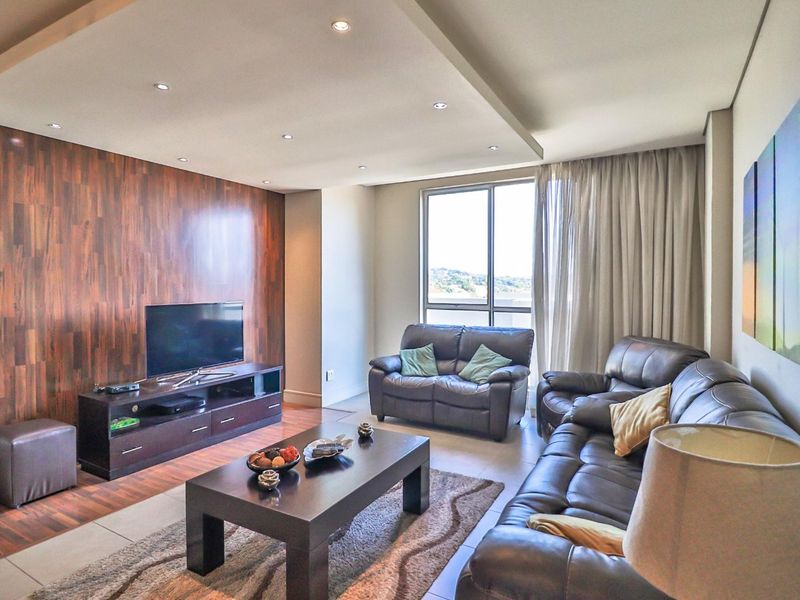 A well-maintained living room featuring a modern wood-paneled feature wall, recessed ceiling lighting, and large windows providing natural light.