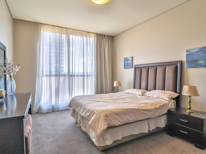 A well-lit bedroom featuring neutral walls, carpeted flooring, and a large floor-to-ceiling window that allows for ample natural light.