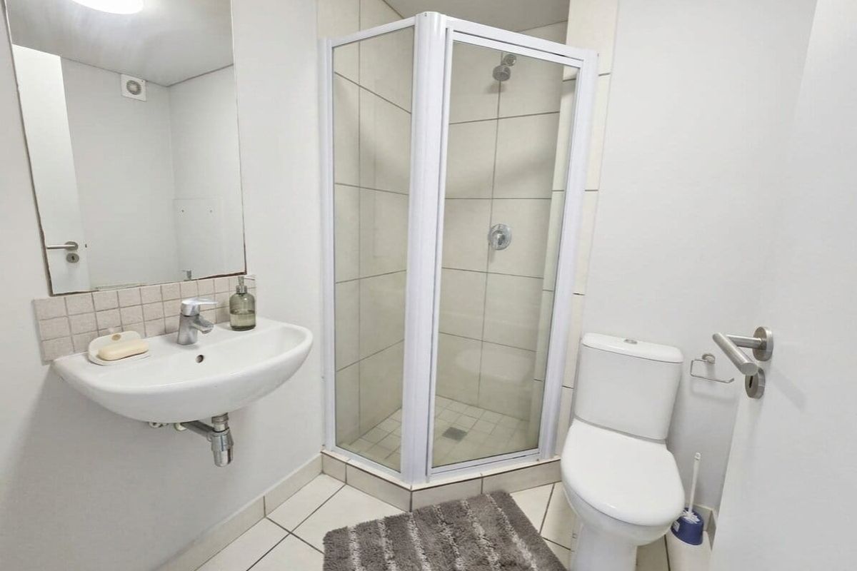 A functional bathroom featuring a corner shower enclosure, wall-mounted basin with mirror, and a standard toilet with tiled flooring.