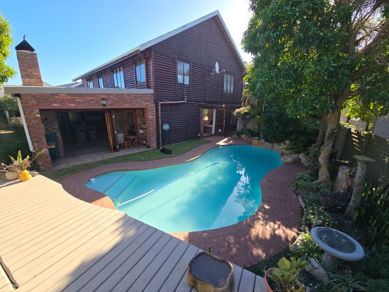 A rear garden view featuring a swimming pool, brick-paved surround, timber decking, and a multi-story log-style home with a covered patio area.