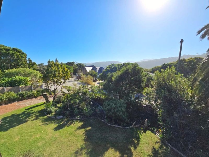 Elevated view overlooking a lush green garden with mature trees and a scenic mountain backdrop under a clear blue sky.