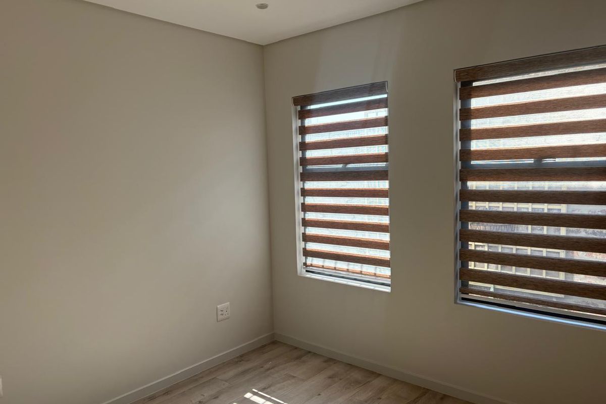 A bright, empty bedroom featuring modern laminate flooring, recessed downlights, and dual-tone roller blinds on the windows.