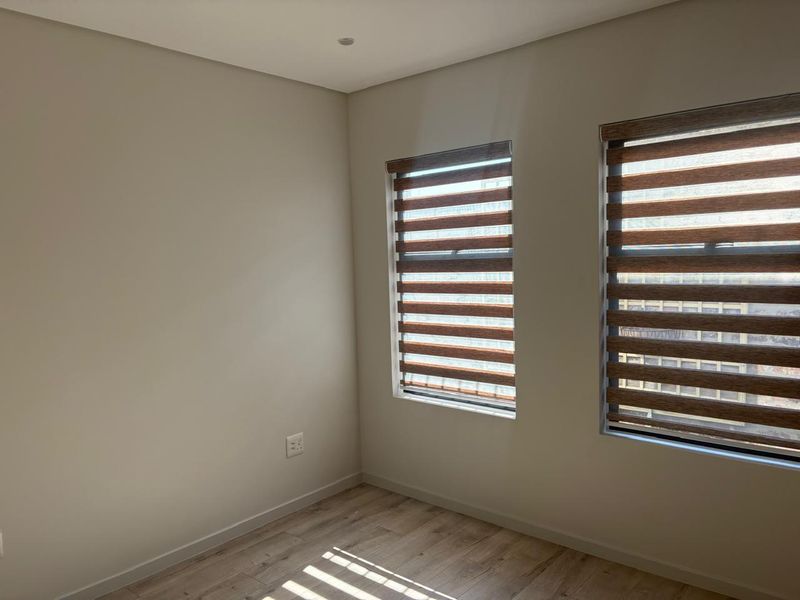 A bright, empty bedroom featuring modern laminate flooring, recessed downlights, and dual-tone roller blinds on the windows.