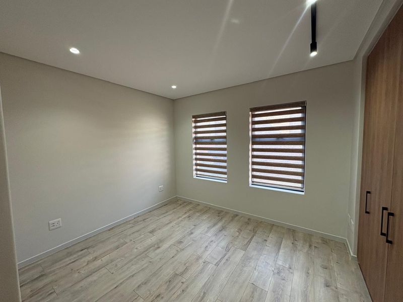 A bright, modern bedroom featuring light wood-look laminate flooring, built-in cupboards, and dual windows with fitted roller blinds.