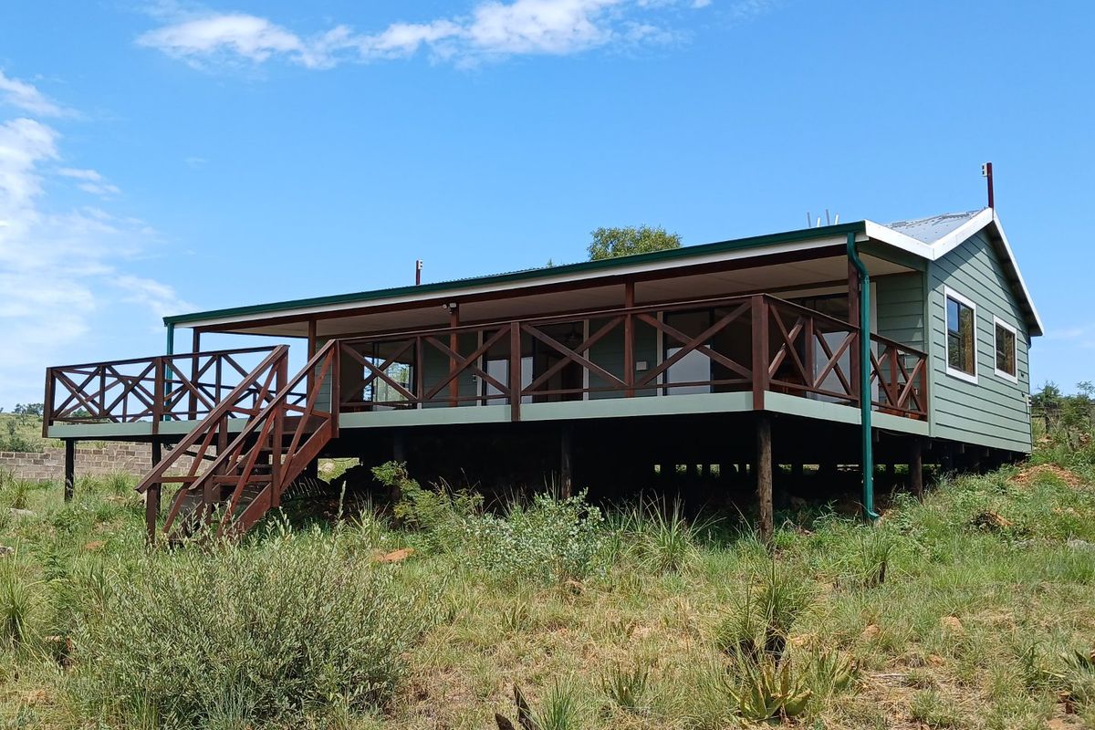 A well-maintained farmhouse featuring a raised wooden deck with balustrades, accessible via an external staircase, set within a natural landscape.