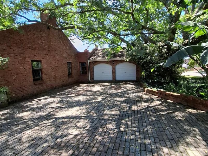Front view of a brick house featuring a spacious brick-paved driveway and a double garage, shaded by mature trees.