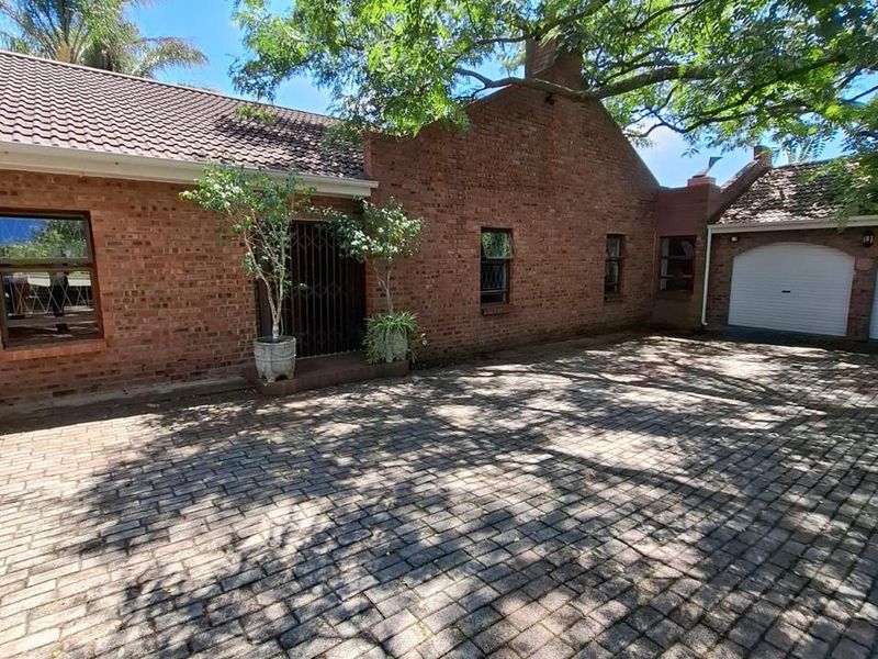 A charming face-brick house exterior featuring a spacious paved driveway, a single garage, and mature trees providing shade to the front entrance.