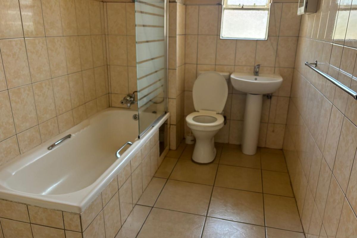 A dated bathroom featuring a bathtub with a glass screen, pedestal basin, and toilet, finished with neutral ceramic wall and floor tiles.