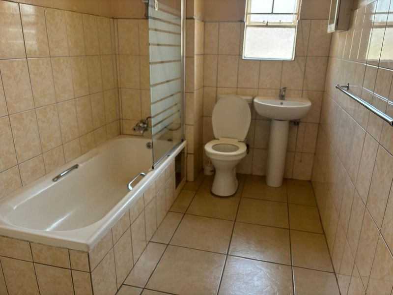 A dated bathroom featuring a bathtub with a glass screen, pedestal basin, and toilet, finished with neutral ceramic wall and floor tiles.