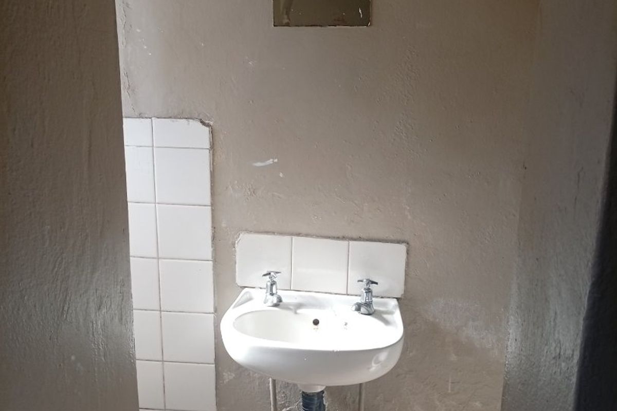 A small bathroom area featuring a wall-mounted basin with exposed plumbing and basic tiled splashback, requiring significant cosmetic renovation and finish improvements.