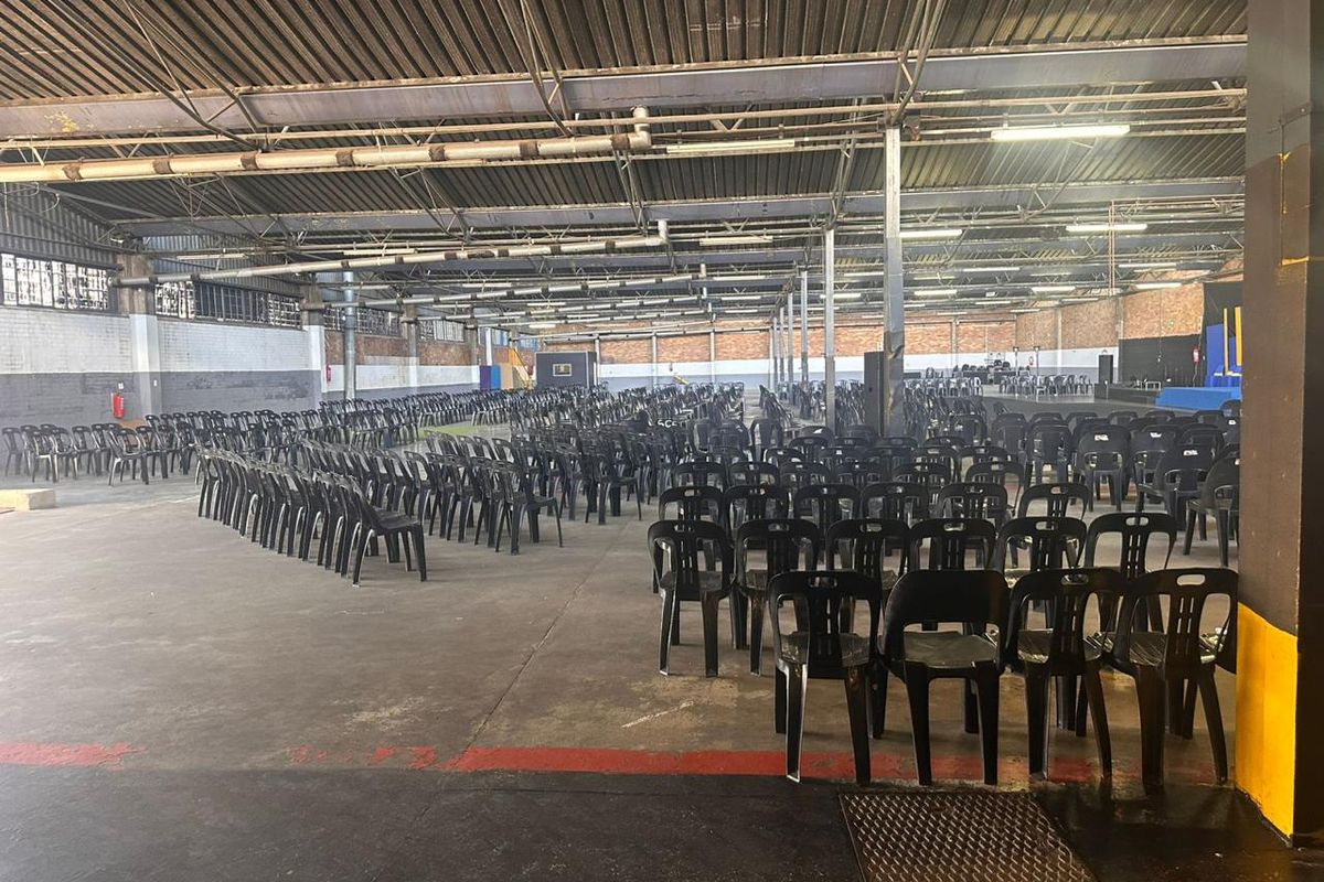 Large industrial warehouse space featuring high exposed steel roof trusses, concrete flooring, and brick masonry walls, currently set up for event seating.